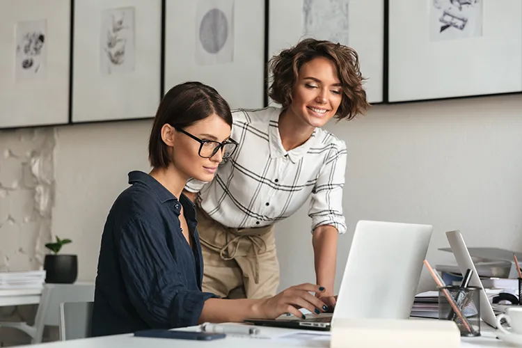 Two women work together at a desk with a laptop. One types while the other stands, smiling at the screen—perhaps discussing how Google reviews do more than build trust—they directly boost your business’s search engine ranking. Artwork decorates the wall behind them.