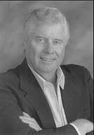 Black and white portrait of an older man with light hair, wearing a suit jacket and collared shirt, smiling with arms crossed.